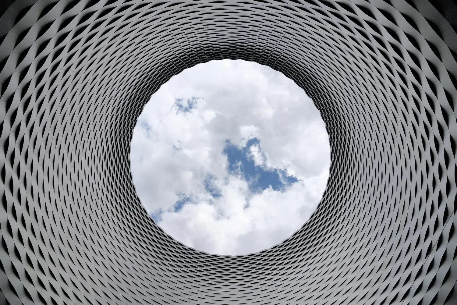 Ein Blick nach oben durch eine große, kreisförmige Öffnung in einer gemusterten Struktur, der den Himmel und die Wolken darüber freigibt, von Tweak.ist